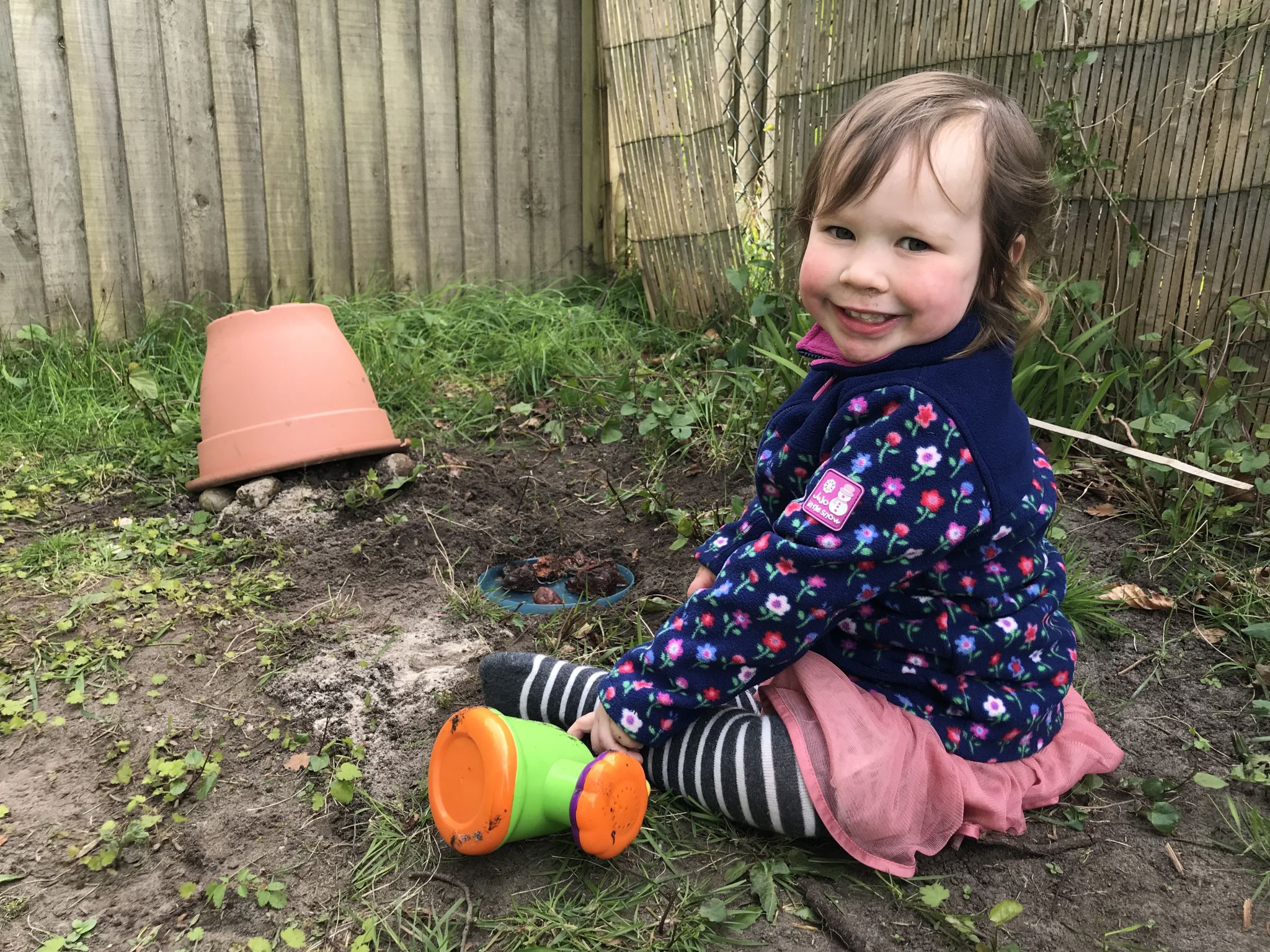 a small child sat in the mud with a homemade toads house out of a pot and rocks