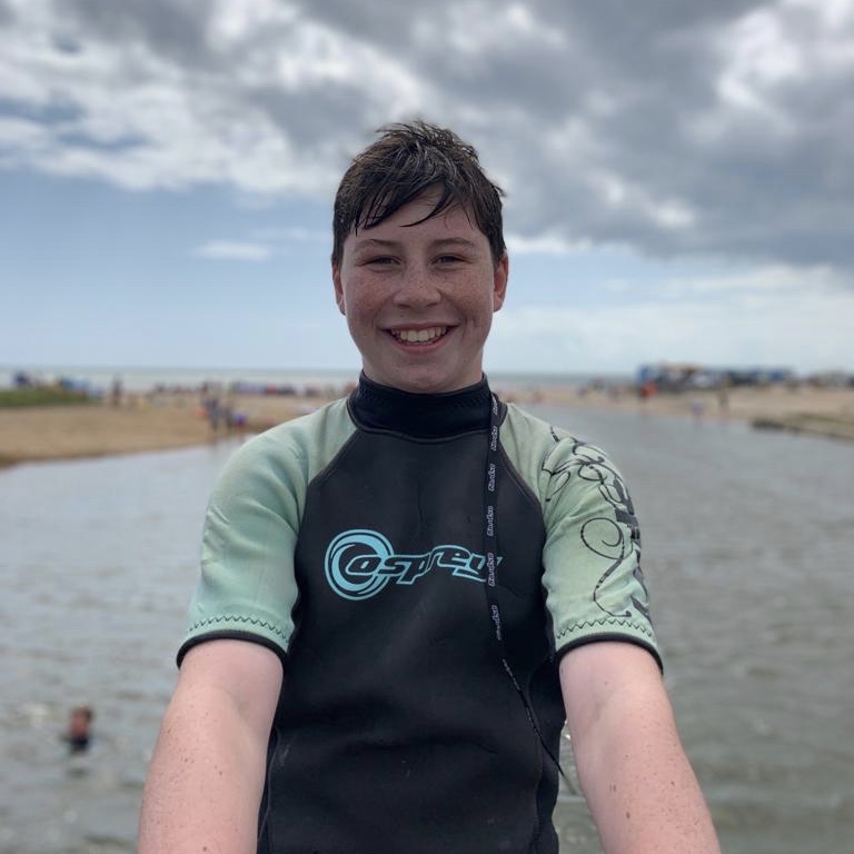 A boy in a wet suit grinning by the sea