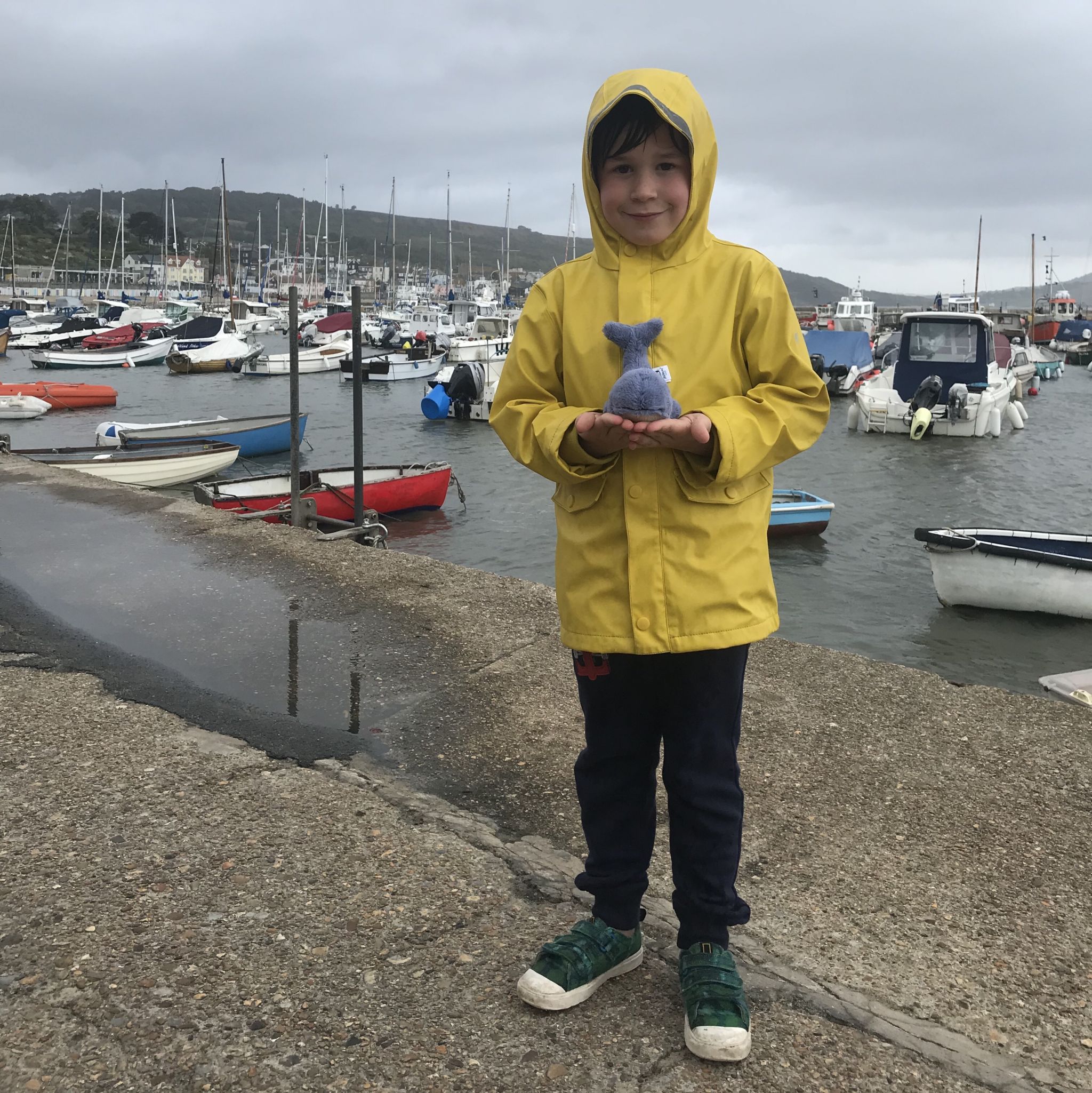 A boy in a yellow fishermans coat holding a toy whale by the harbour