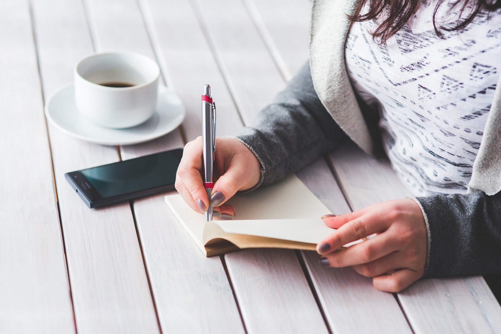 a lady writing down her thoughts in a notebook with a hot drink next to her 