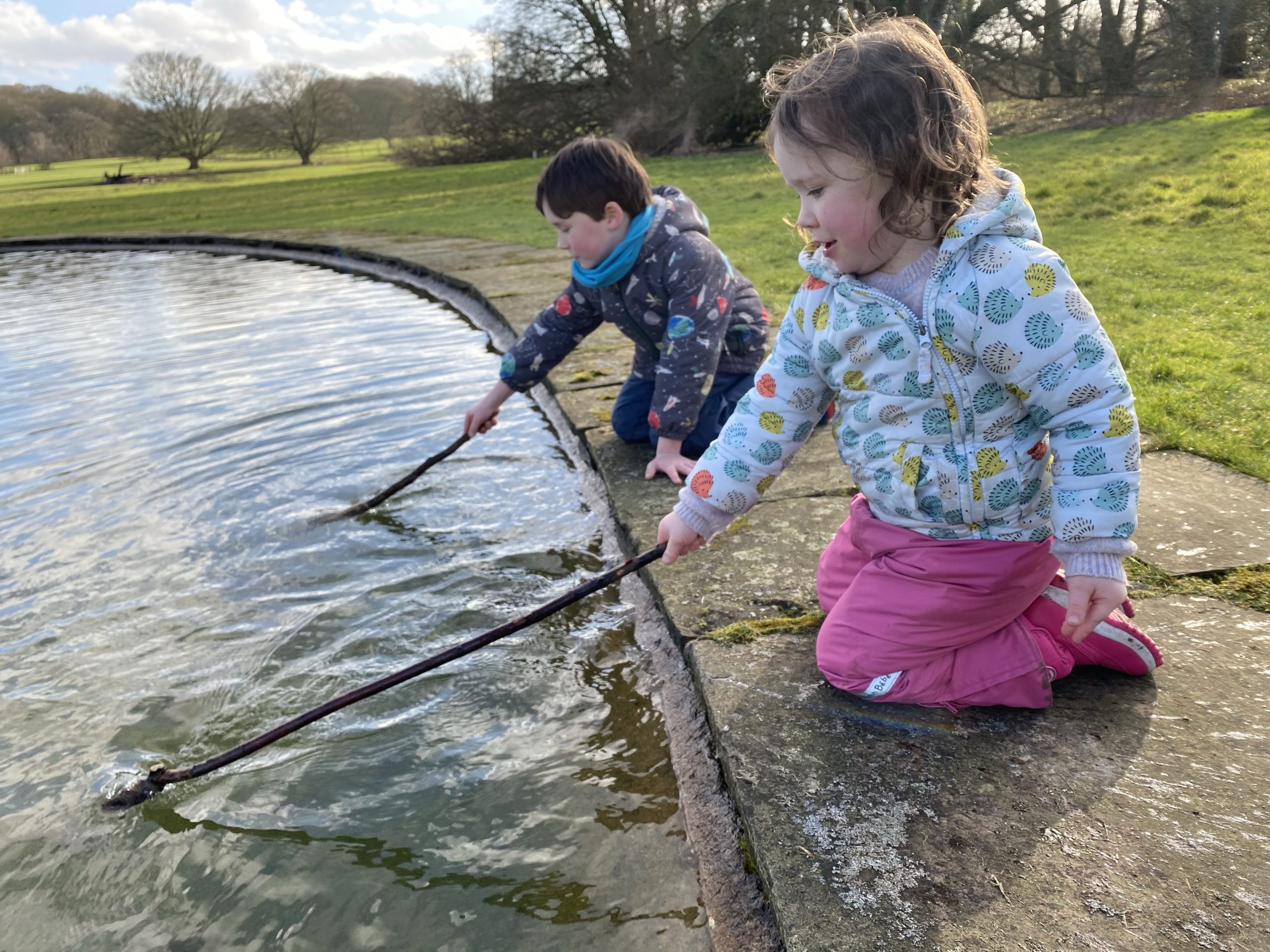 Two small children playing by a pond with sticks 