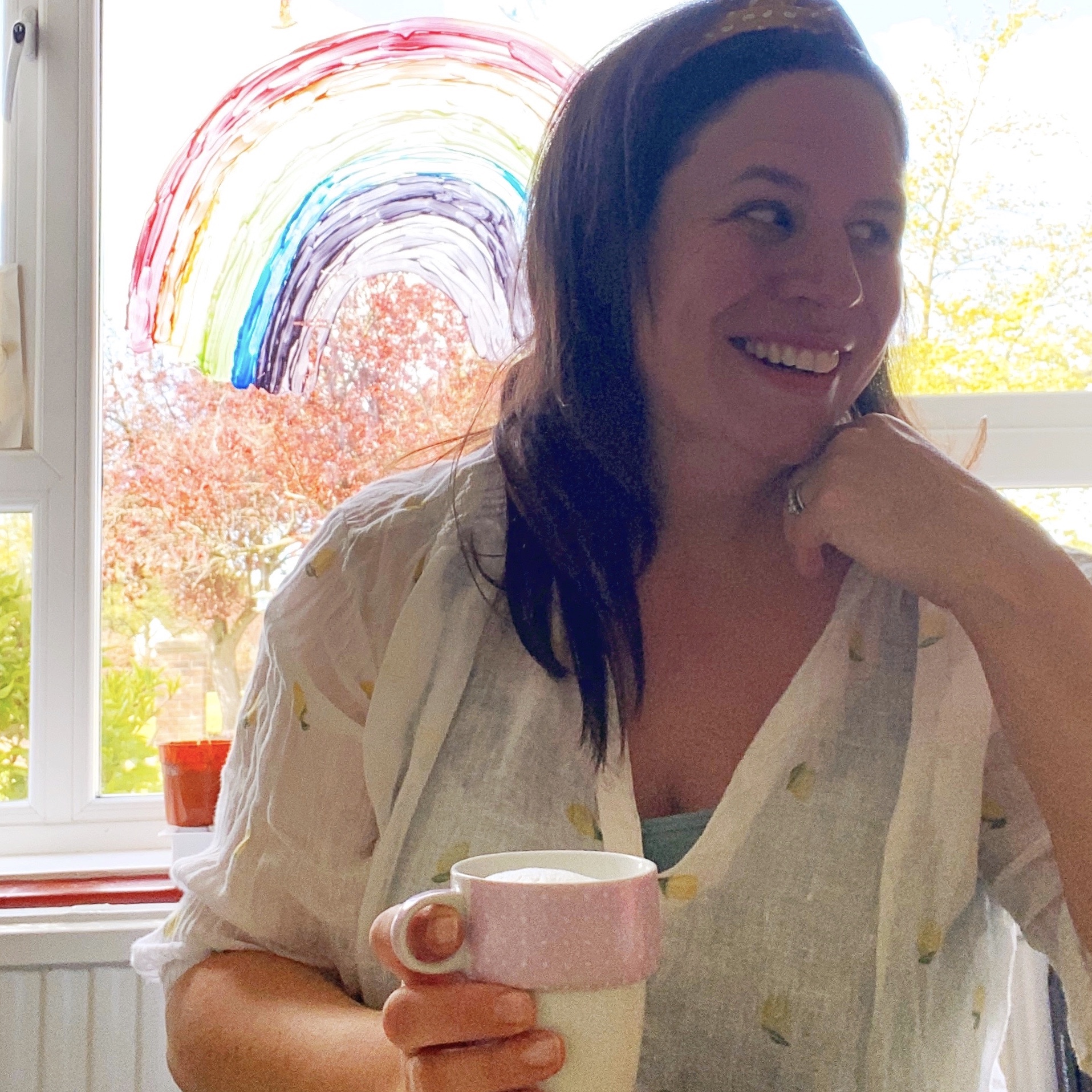 a woman drinking coffee and smiling by her stay safe indoors rainbow on her window 